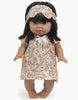 A Minikane | Nightgown & Sleeping Mask in Bunny Print with dark hair and brown skin wearing a floral dress and matching headband. The 13" doll's dress features a colorful pattern with flowers and leaves. The doll stands upright on a plain background, showcasing its charming outfit.