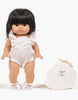 A Minikane Gordis doll with black hair wearing a Minikane Doll Clothing | Lou Romper in Cream Cotton and white sandals stands next to a small off-white drawstring bag labeled "minikane." The doll has a neutral expression, and both items are set against a plain white background.