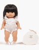 A Minikane Gordis doll with straight black hair wears the Minikane 13" Doll Clothing Lou Retro Romper in Cotton Gauze - Cream and white sandals, standing next to a white double gauze drawstring bag. The background is plain white.