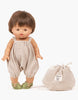 A Minikane Gordis doll with short brown hair, wearing a light beige romper and green sandals, stands next to a matching beige drawstring bag labeled "Minikane Doll Clothing | Kim Bloomers in Raw Cotton." The background is white.