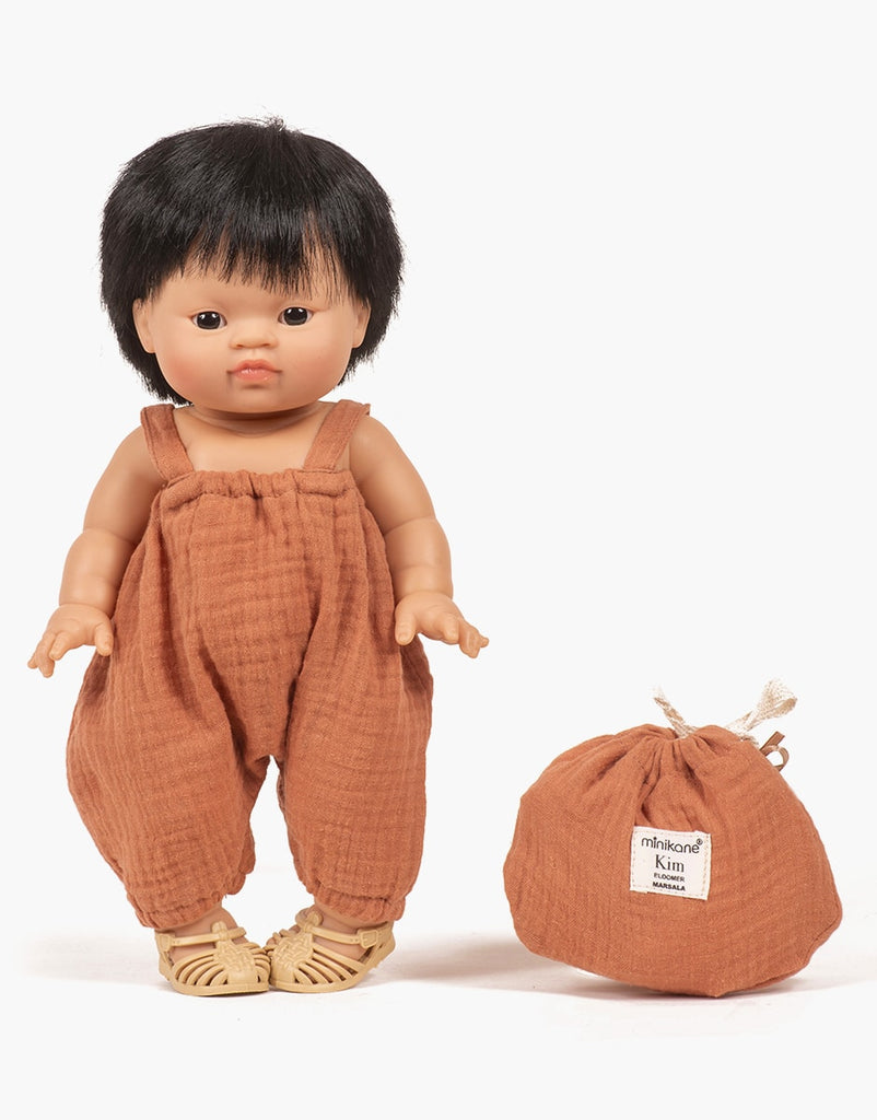 A doll with short black hair wears a brown romper and cream-colored woven shoes, crafted from double gauze fabric. Beside the doll, there is a matching brown cloth bag with a label that says "Minikane Doll Clothing | Kim Bloomers in Marsala." The background is plain white. Part of the exquisite Minikane Gordis dolls collection, this set also features adorable Kim bloomers for mix-and-match styling.