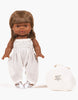 A Minikane Gordis doll with brown hair and dark skin stands upright, dressed in a Minikane Doll Clothing | Bonnie Jumpsuit in Cream crafted from double gauze. Next to the doll is a white fabric bag with text on it. The background is plain white.