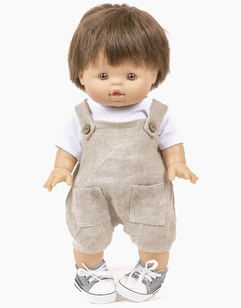 A Minikane Gordis doll with light skin, straight brown hair cut in a short bob, and brown eyes. The doll is wearing the Minikane Doll Clothing | Antonin Overalls Set Doll Clothing, which consists of linen overalls, a white t-shirt, and white sneakers with grey accents. It stands upright against a plain white background.