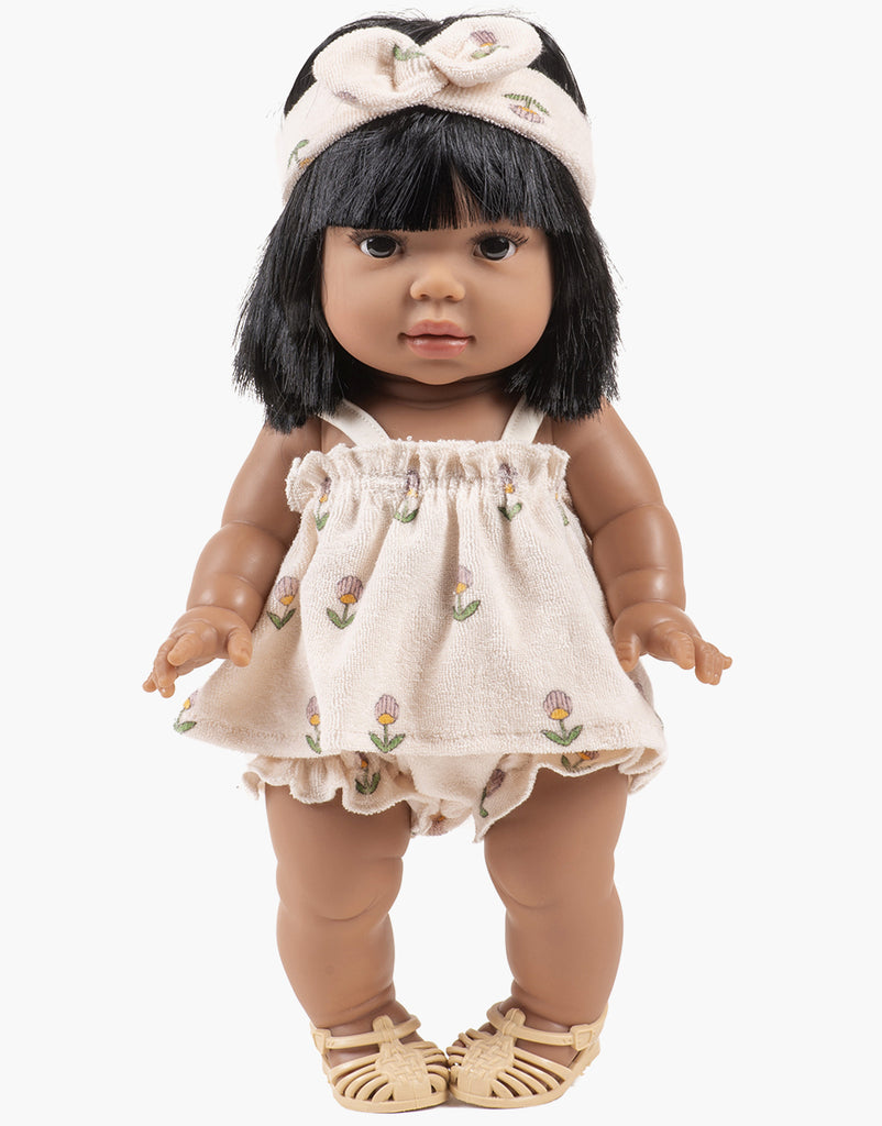 The Minikane Doll, standing in brown sandals, wears the Mila Soft Flowers Set in Terrycloth by Emile & Ida with a cream headband and short black hair, exuding French charm. Hand washable for easy care.