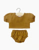 A brown baby outfit consisting of a short-sleeved top and elastic waistband bottoms is displayed on a white background. The Minikane Clothing | Charlotte Balloon Sleeve Shirt and Briefs set, perfect for Minikane Gordis dolls, is hanging on a small wooden hanger with a metal hook.