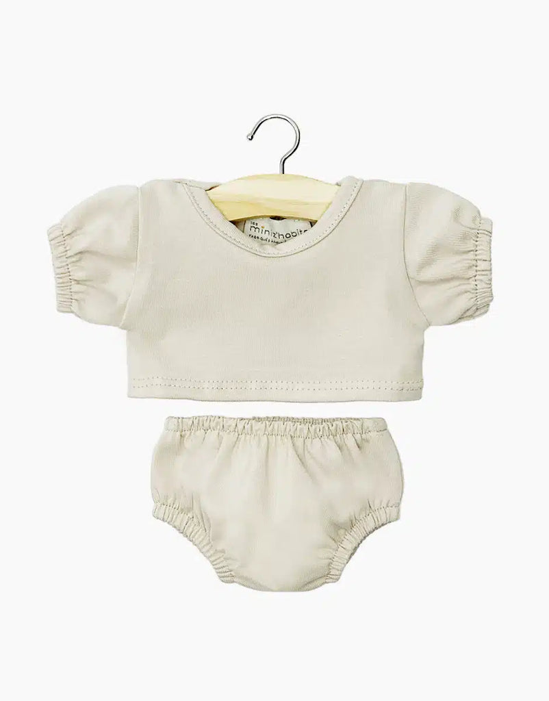 A beige baby outfit on a hanger, featuring a balloon-sleeved t-shirt with gathered sleeves and matching elastic waistband shorts. Both pieces are simple and unadorned, made from soft fabric. The hanger is wooden with a metal hook, perfect for Minikane Doll Clothing | Charlotte Balloon Sleeve T-Shirt and Briefs Set in Linen Ecru displays.