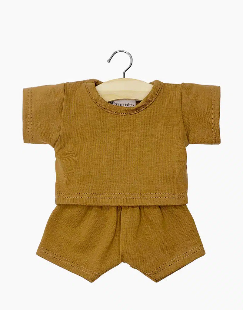 A small brown baby outfit, perfect for 13 & 14 inch doll clothes, features a short-sleeve top and shorts displayed on a wooden hanger with a metal hook. Made of soft fabric, this Minikane Doll Clothing | Vito T-Shirt and Shorts Set in Camel boasts simple, clean stitching.