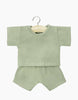 A light green baby outfit, perfect for Minikane Gordis dolls, hangs on a small wooden hanger against a white background. The outfit consists of a short-sleeved shirt akin to the Minikane Doll Clothing | Vito T-Shirt and Shorts Set in Green and matching shorts, both featuring subtle stitched detailing on the edges.