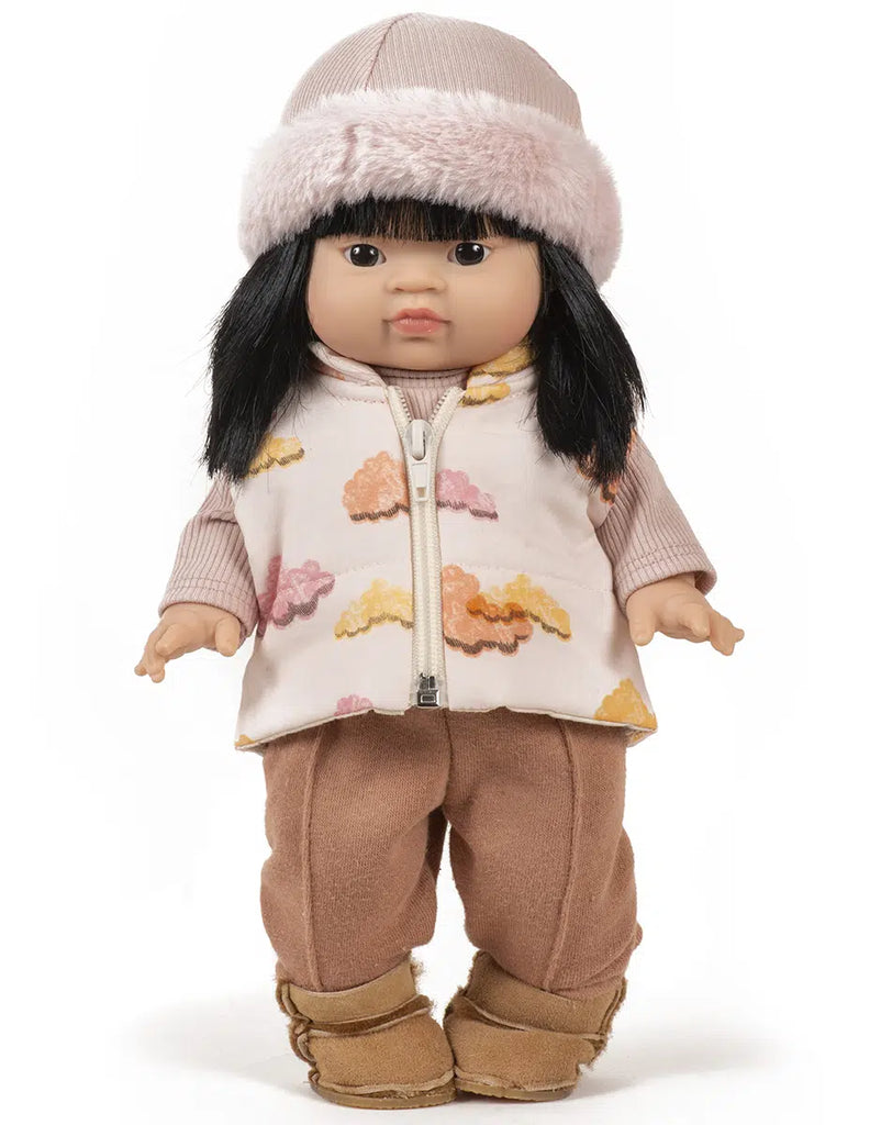 The Minikane doll, featuring long black hair, wears a pink furry hat, petal ribbed knit undershirt, cloud-printed vest, tan pants with brown boots. The set's French craftsmanship and neutral expression amplify its timeless appeal.