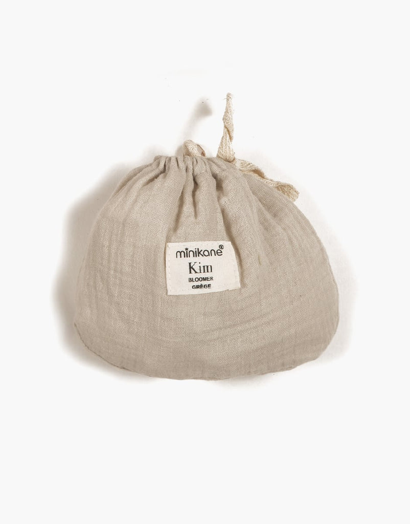 A beige drawstring bag with textured double gauze fabric. A rectangular white label on the front reads "Minikane Doll Clothing | Kim Bloomers in Raw Cotton". Ideal for storing Minikane Gordis dolls, the bag is cinched at the top with a tied string closure.