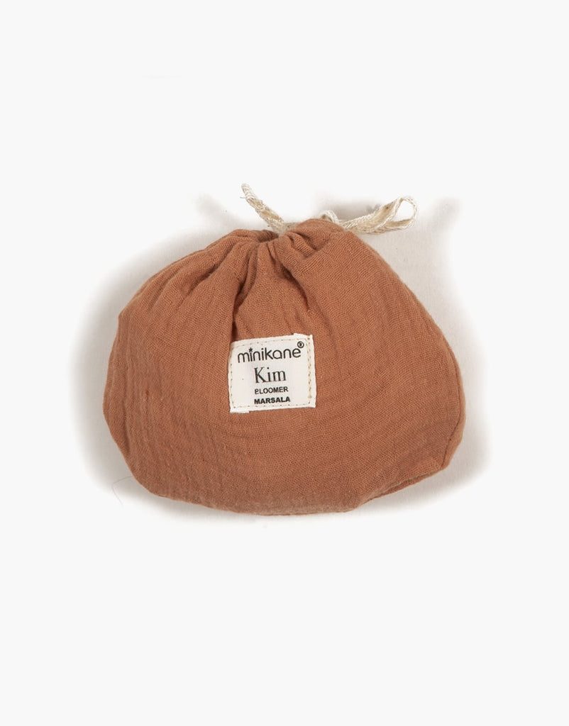 A small, brown fabric pouch with a drawstring closure is shown against a white background. The pouch, ideal for Minikane Gordis dolls, features a white label with black text that reads "Minikane Doll Clothing | Kim Bloomers in Marsala" in two lines and is made from soft double gauze material.