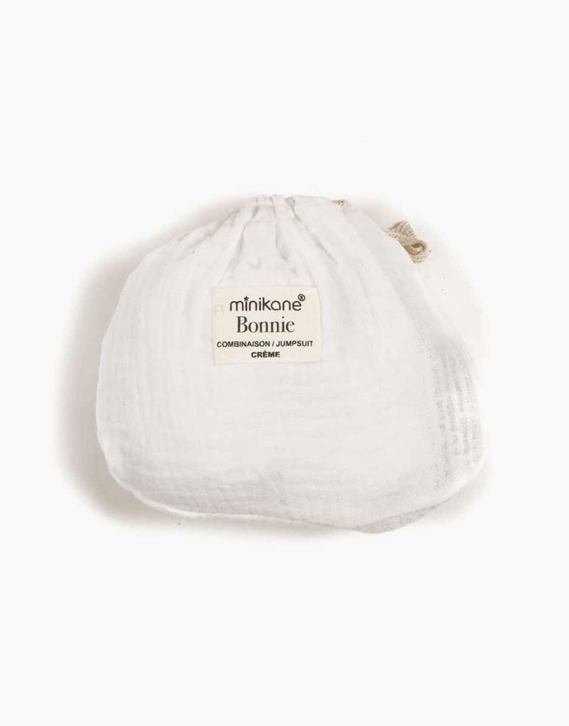 A white, round fabric pouch labeled "Minikane Doll Clothing | Bonnie Jumpsuit in Cream" containing a cream-colored Long Bonnie jumpsuit crafted from double gauze. The garment is described as a "COMBINAISON/JUMPSUIT" on the label and fits Minikane Gordis dolls. The pouch has a drawstring closure at the top.