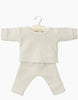 A set of light beige, waffle-knit baby clothing hangs on a wooden hanger. The set, reminiscent of cozy Minikane Doll Clothing | Morgan Pajamas in Honeycomb Linen Knit, includes a long-sleeve top and matching pants with visible textured patterns. Perfect for Minikane Gordis dolls or similarly sized 13 & 14" companions. The background is plain white.