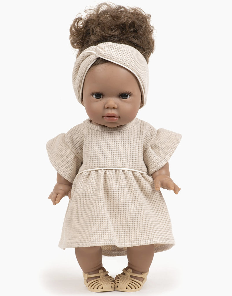 A Minikane Gordis doll with curly brown hair, wearing a beige headband and a matching Minikane Doll Clothing | Linen Dress and Headband Set with flared sleeves, stands upright. The doll has a neutral expression and is also wearing beige shoes.
