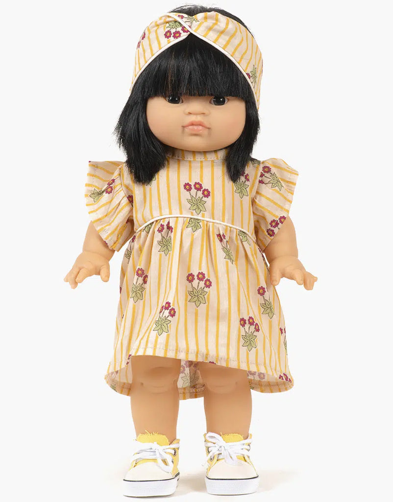 A Minikane doll with black hair wears the Daisy Dress and a matching crossed headband featuring floral patterns. Designed in France, it comes with hand washable white sneakers.