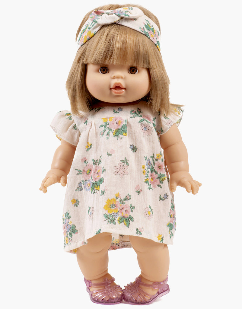 A Minikane doll with short blonde hair dons an Emile & Ida pink spring dress and matching headband in cotton, paired with purple sandals. Designed in France, this charming doll is hand washable for easy care.