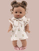 A Minikane doll wears the Emile & Ida Emilia Dress in Soft Terrycloth - Flower, a cream outfit with small prints, and pink sandals. The brown-skinned doll features curly hair in two buns and stands against a beige background. Designed in France.