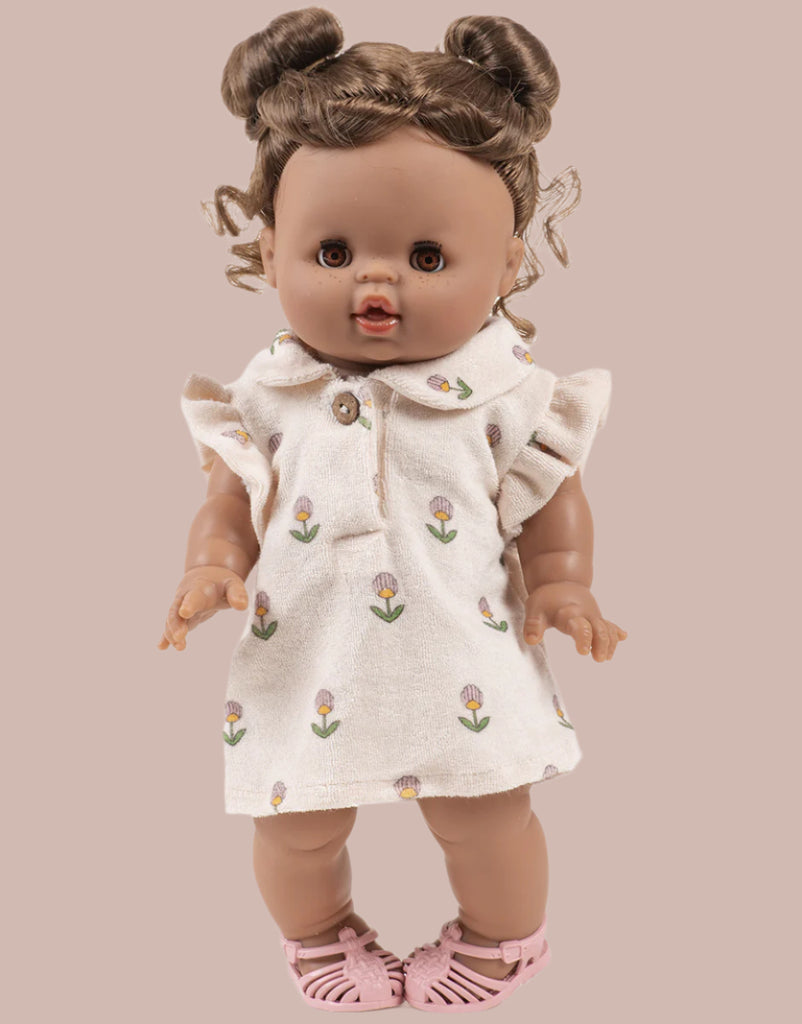 A Minikane doll wears the Emile & Ida Emilia Dress in Soft Terrycloth - Flower, a cream outfit with small prints, and pink sandals. The brown-skinned doll features curly hair in two buns and stands against a beige background. Designed in France.