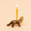 A Mink Cake Topper figurine featuring a cute mink wearing a tiny party hat and carrying a yellow birthday candle on its back, displayed against a plain beige background.