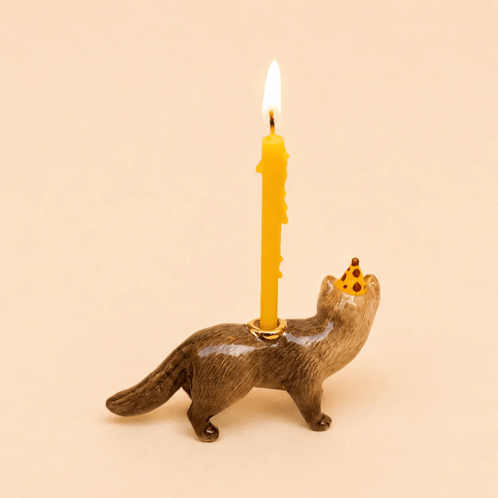 A Mink Cake Topper figurine featuring a cute mink wearing a tiny party hat and carrying a yellow birthday candle on its back, displayed against a plain beige background.