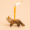 The Mink Cake Topper - Final Sale is a charming porcelain figurine of a brown animal wearing a yellow party hat and holding an upright lit yellow keepsake birthday candle on its back, making it an adorable cake topper for any celebration.