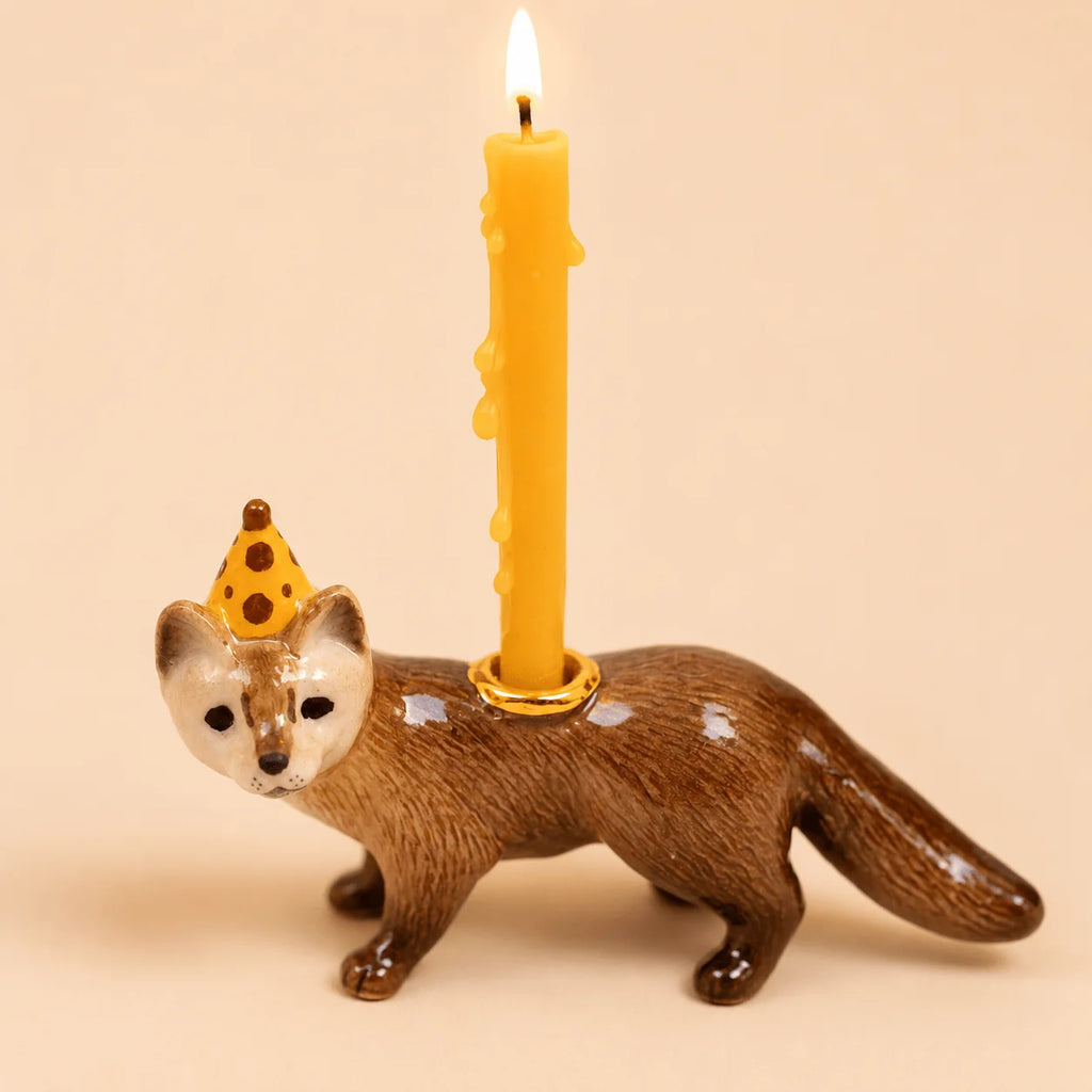 The Mink Cake Topper - Final Sale features a ceramic figurine of a brown weasel sporting a yellow party hat. Designed to securely hold a birthday candle on its back, the topper showcases melted wax gently dripping down the lit candle, set against a soft beige background.