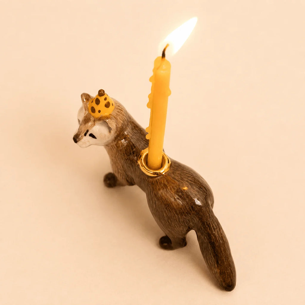 The Mink Cake Topper – Final Sale features a charming ceramic fox figurine with a yellow candle holder on its back. The fox wears a small yellow crown adorned with brown dots, and the candle holder displays a lit yellow birthday candle. This unique porcelain keepsake is perfect for topping cakes at special celebrations.