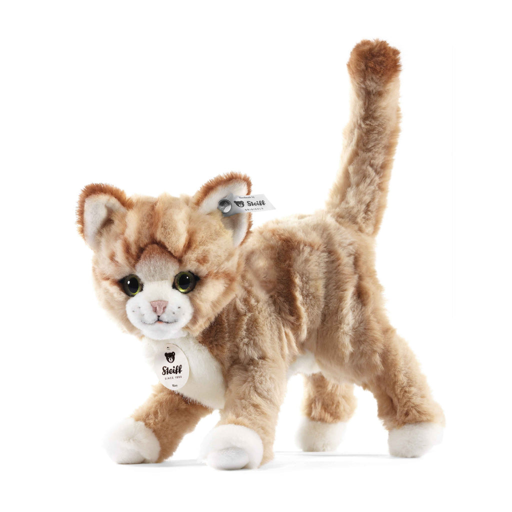 The Steiff Mizzy Cat is a soft, light brown plush cat with white paws and face, green eyes, and an upright tail. It features Steiff tags on its ear and chest, standing on all four legs against a white background.