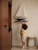 A young child in a striped outfit and yellow vest reaches up toward the Little Forest Animals Silk Rug — The Regatta Series — The Endeavour, beautifully displayed on a shelf in a room with striped wallpaper and wooden furniture.