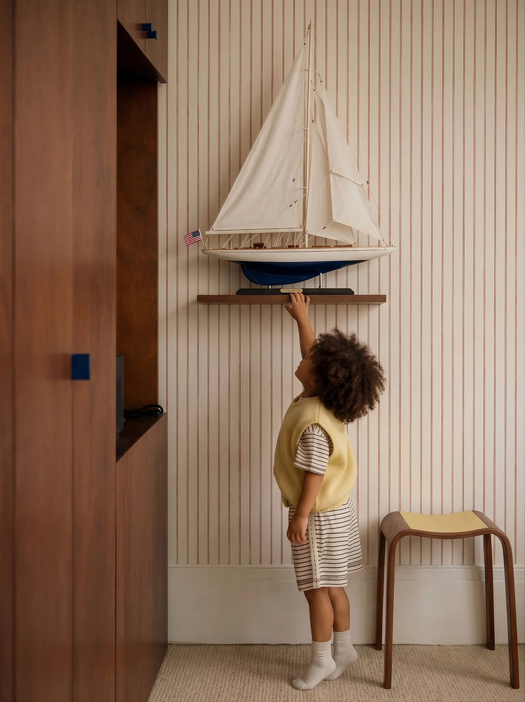 A young child in a striped outfit and yellow vest reaches up toward the Little Forest Animals Silk Rug — The Regatta Series — The Endeavour, beautifully displayed on a shelf in a room with striped wallpaper and wooden furniture.