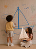 Two children create a nautical scene; one draws a large blue sailboat on the wall with crayons, while the other sits holding a handcrafted sailing yacht, nestled atop the Little Forest Animals Silk Rug — The Regatta Series — The Endeavour, surrounded by colorful drawings of a house, sun, clouds, and a tree.