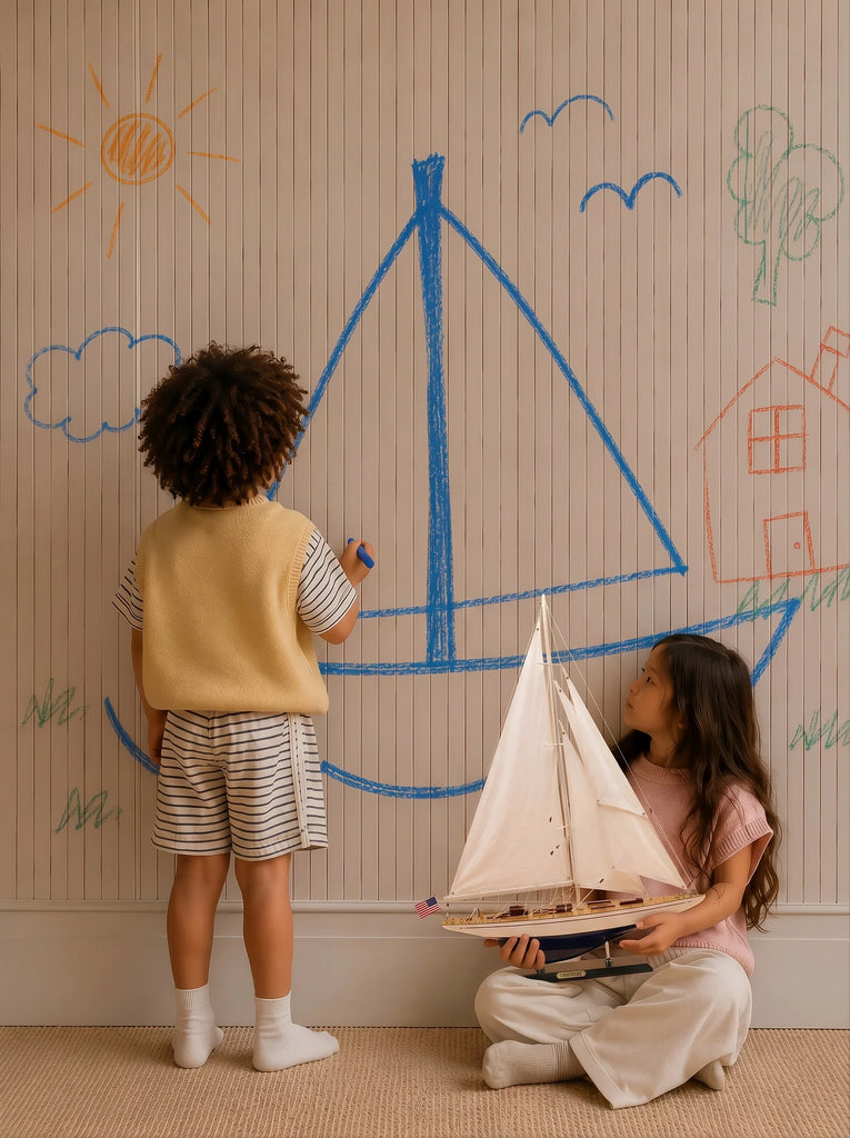 Two children create a nautical scene; one draws a large blue sailboat on the wall with crayons, while the other sits holding a handcrafted sailing yacht, nestled atop the Little Forest Animals Silk Rug — The Regatta Series — The Endeavour, surrounded by colorful drawings of a house, sun, clouds, and a tree.