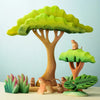 A whimsical scene features the Bumbu Wooden Monkey Set in a colorful jungle setting with two large, vibrant trees and lush tropical plants. Inspired by Montessori and Waldorf principles, one eco-friendly monkey perches on a tree while others gather around the base against a clear blue background.