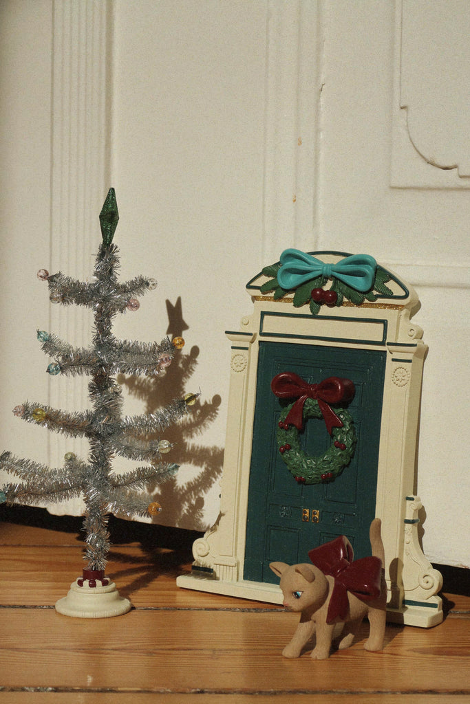 A Konges Sloejd Christmas Door featuring a miniature decorative door with a wreath and bow, accompanied by a small toy cat with a bow and a tinsel tree.