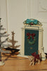 A Konges Sloejd Christmas Door featuring a festive wreath and blue bow, displayed on a wooden surface next to a small tinsel tree and a toy kitten wearing a red bow as a festive elf accessory.