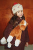 A young girl wearing a floral beanie smiles while holding a cuddly Konges Sloejd Fiona Horse.