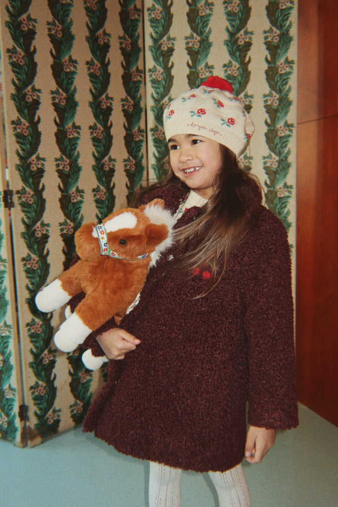 A smiling girl wearing a beanie and coat holds a cuddly Konges Sloejd Fiona Horse toy.