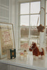 A soft Konges Sloejd Fiona Horse toy stands on a windowsill next to a small lamp, a snow globe, and a framed art poster.