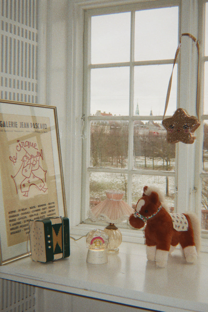 A soft Konges Sloejd Fiona Horse toy stands on a windowsill next to a small lamp, a snow globe, and a framed art poster.