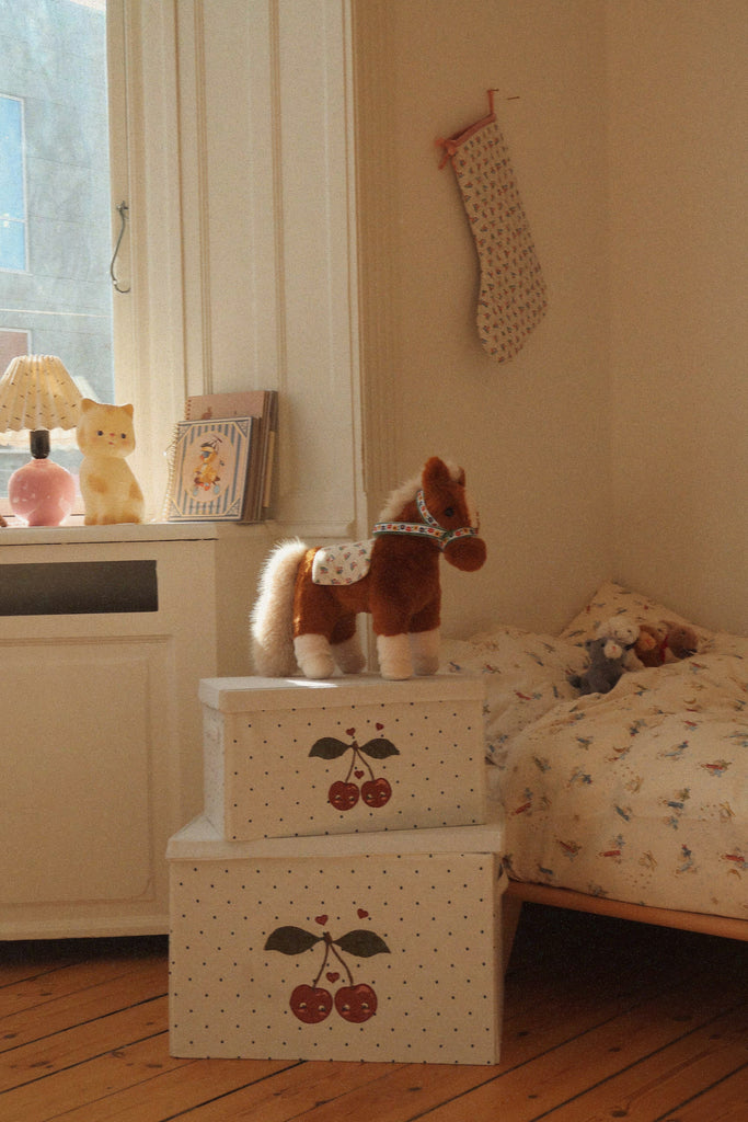 A brown, soft Konges Sloejd Fiona Horse toy with a white saddle stands on a stack of two white storage boxes decorated with cherry illustrations.
