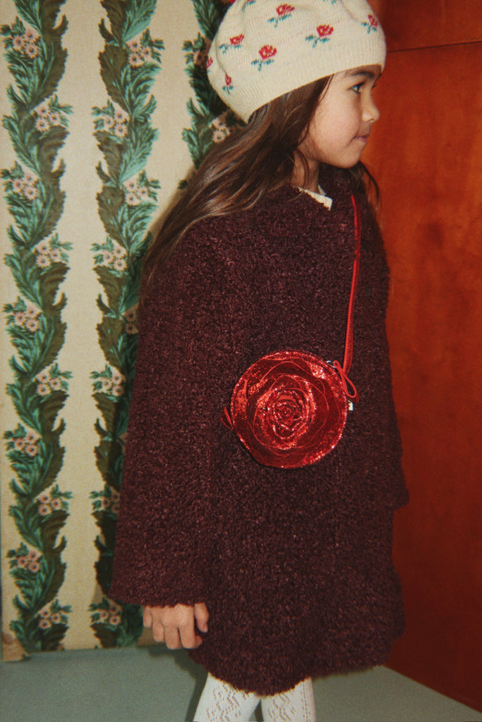 A child wearing a cream-colored beanie and a dark textured coat carries a round, red Konges Sloejd Rose Shoulder Bag with a slim shoulder strap.
