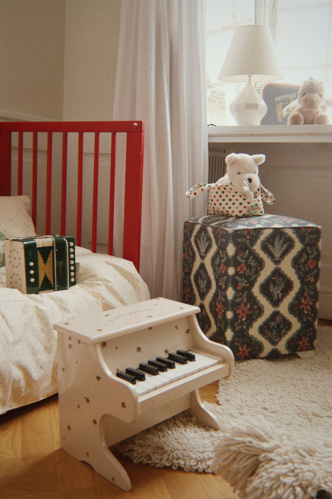 A Konges Sloejd Accordian 10-key button toy resting on a child’s bed next to a small wooden piano.