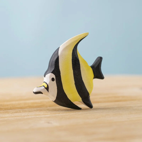 The Bumbu Handmade Wooden Moorish Idol Fish, featuring hand-painted black, white, and yellow stripes, sits on a light wooden surface against a pale blue background.