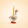 The Mother Goose Cake Topper, a handcrafted ceramic figurine wearing a brown hat and carrying a lit yellow candle on its back, makes a charming birthday decoration against a plain beige background.