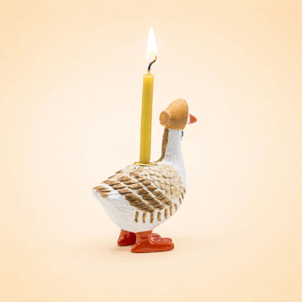 The Mother Goose Cake Topper, a handcrafted ceramic figurine wearing a brown hat and carrying a lit yellow candle on its back, makes a charming birthday decoration against a plain beige background.
