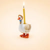 A ceramic “Mother Goose Cake Topper” figurine wearing a small hat holds a lit yellow candle on its back, set against a plain beige background—perfect as a handcrafted birthday decoration or charming collectible keepsake.
