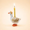 The Mother Goose Cake Topper - Final Sale features a ceramic goose wearing a bonnet and carrying a lit yellow candle on its back. Set against a pale peach background, this charming handcrafted piece makes an adorable birthday decoration or unique collectible keepsake.