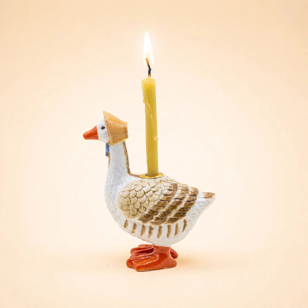 The Mother Goose Cake Topper - Final Sale features a ceramic goose wearing a bonnet and carrying a lit yellow candle on its back. Set against a pale peach background, this charming handcrafted piece makes an adorable birthday decoration or unique collectible keepsake.