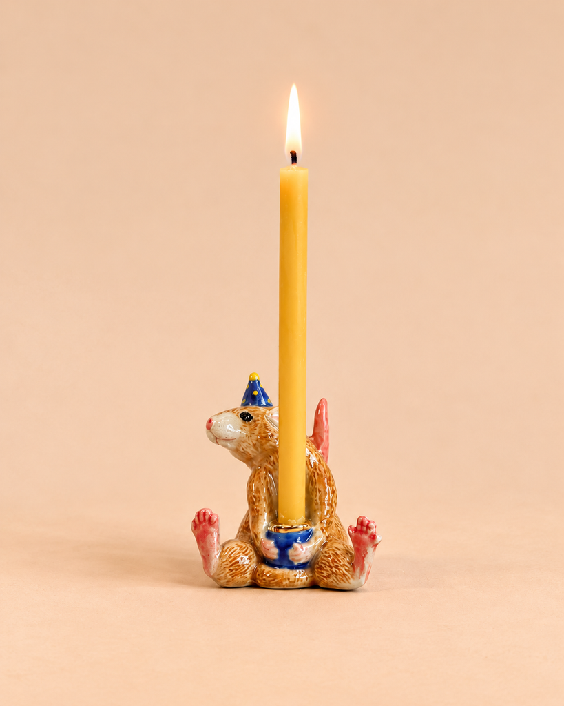 A handcrafted collectible ceramic figurine from the Mouse Cake Topper - Final Sale collection holds a lit yellow candle, depicting a mouse wearing a blue party hat and sitting upright with its legs spread out on a beige background.