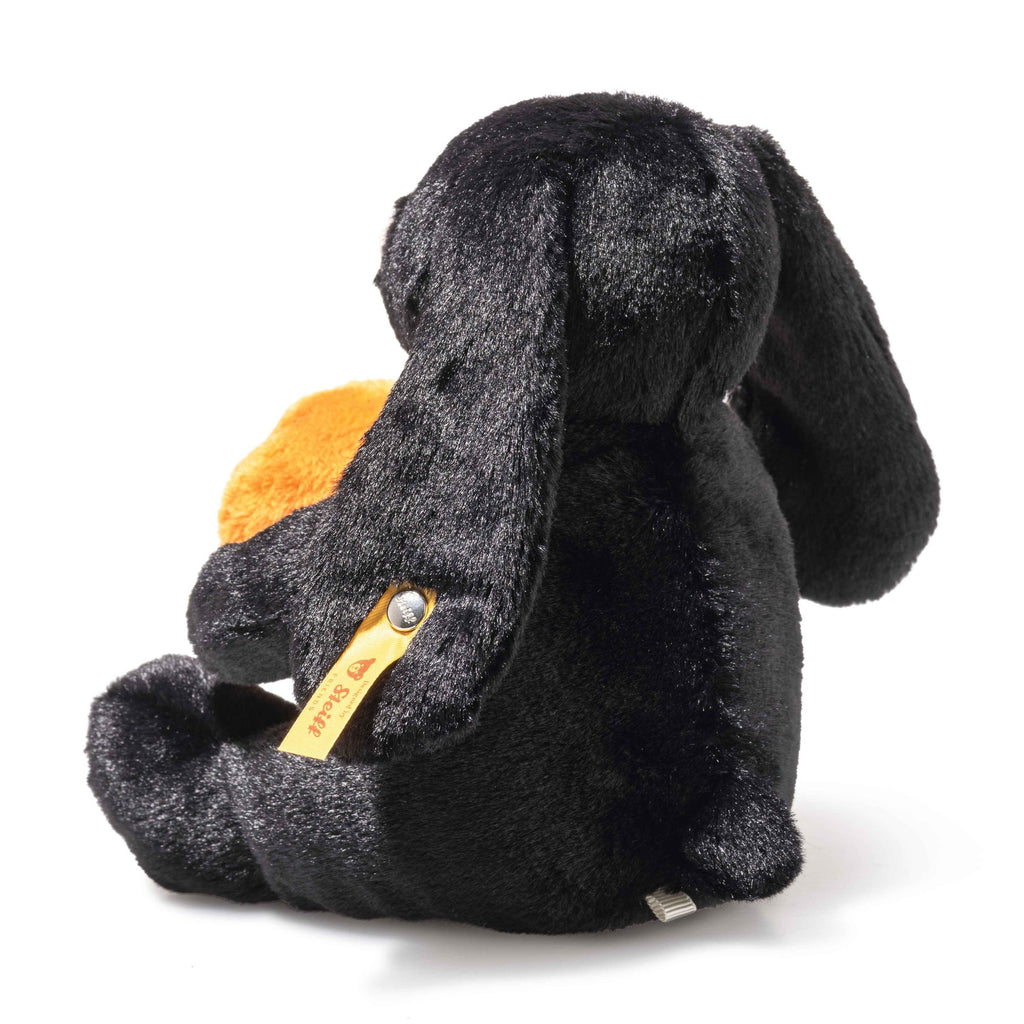 The Steiff Mr. Spice Hoppie Rabbit is a soft black plush with long floppy ears, sitting facing away while holding an orange heart. It features a fuzzy surface and has the signature yellow Steiff Button in Ear tag attached.